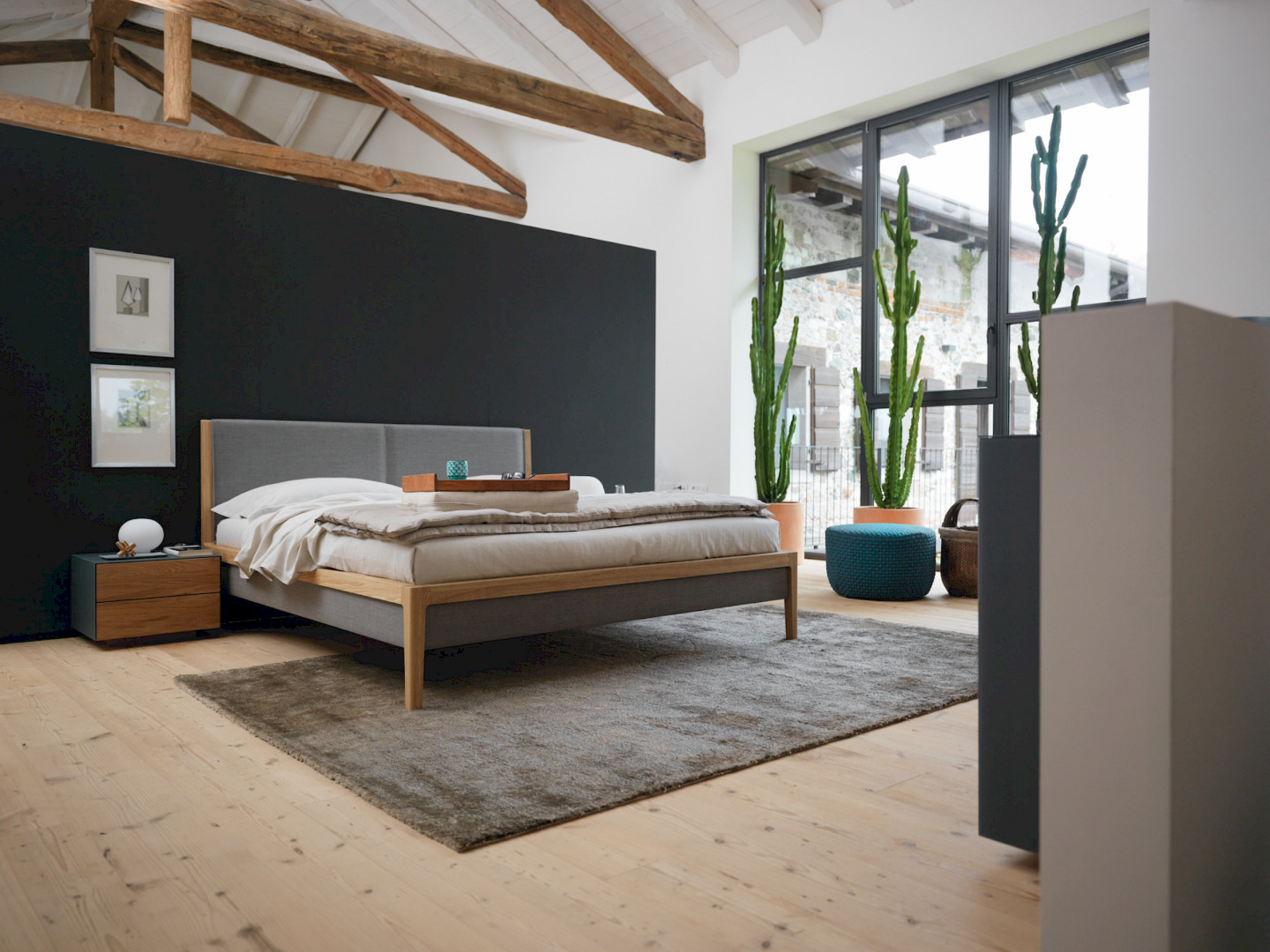 Massivholzbett mit grauer Polsterung in einem loftartigen Raum mit offenem Dachstuhl, Dachbalken und einer großen Fensterfront.