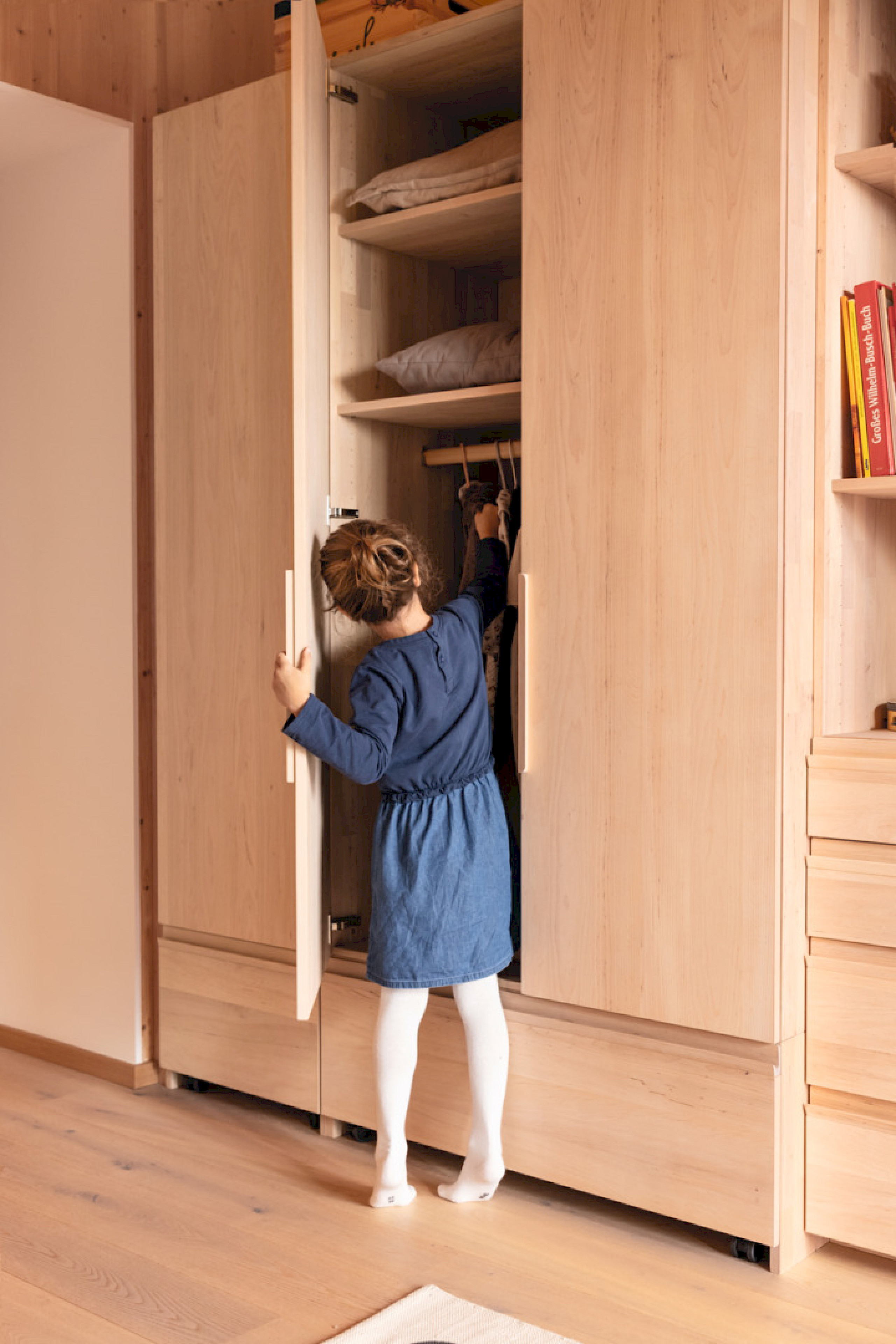 Ein Kind, von hinten aufgenommen, steht vor einem Kleiderschrank aus Holz auf den Zehenspitzen und möchte ein Kleidungsstück herausnehmen